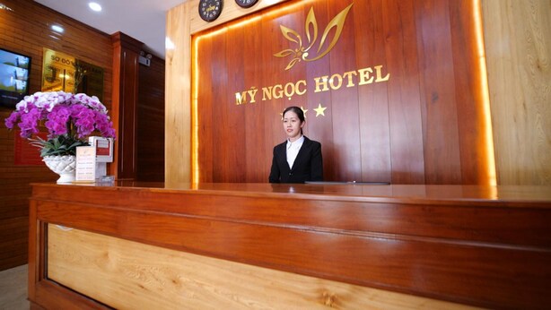 A wooden reception desk at MY NGOC HOTEL with a clock and a floral arrangement.