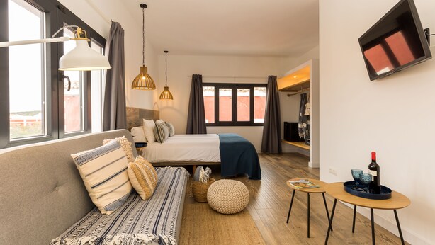A modern hotel room with a sofa, bed, and a small table with a wine bottle. - Pardela Menorca Hotel (Es Castell)