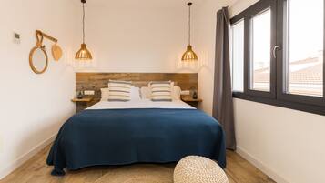 A bedroom with a wooden headboard, a blue bedspread, two bedside lamps, a bedside table, and a large window with curtains.