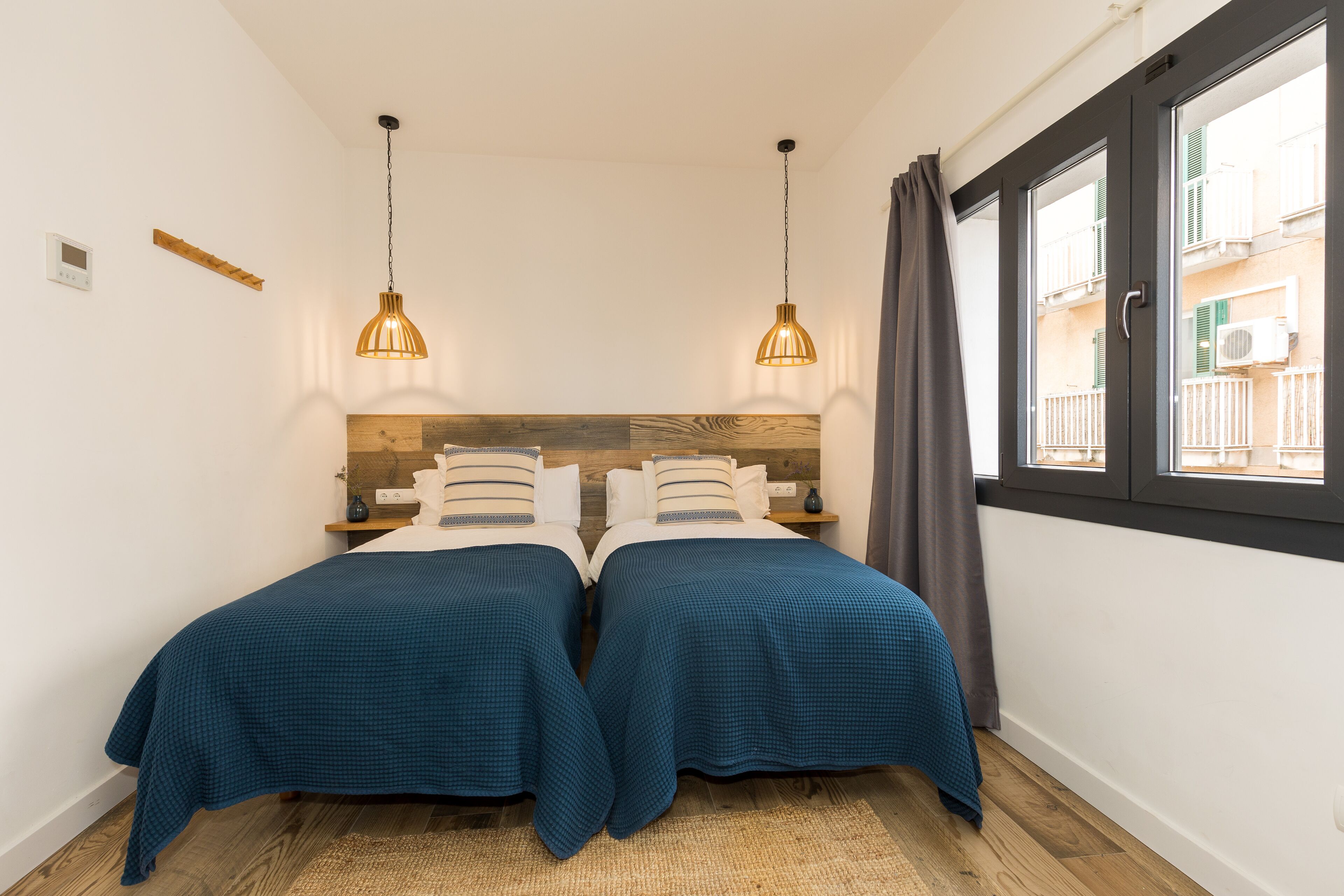 Ein Schlafzimmer mit zwei Betten, einem Holzkopfstück, hängenden Pendelleuchten, einem Fenster mit Blick auf Gebäude und einem kleinen Regal mit Gegenständen.