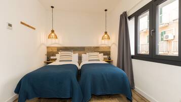 Ein Schlafzimmer mit zwei Betten, einem Holzkopfstück, hängenden Pendelleuchten, einem Fenster mit Blick auf Gebäude und einem kleinen Regal mit Gegenständen.