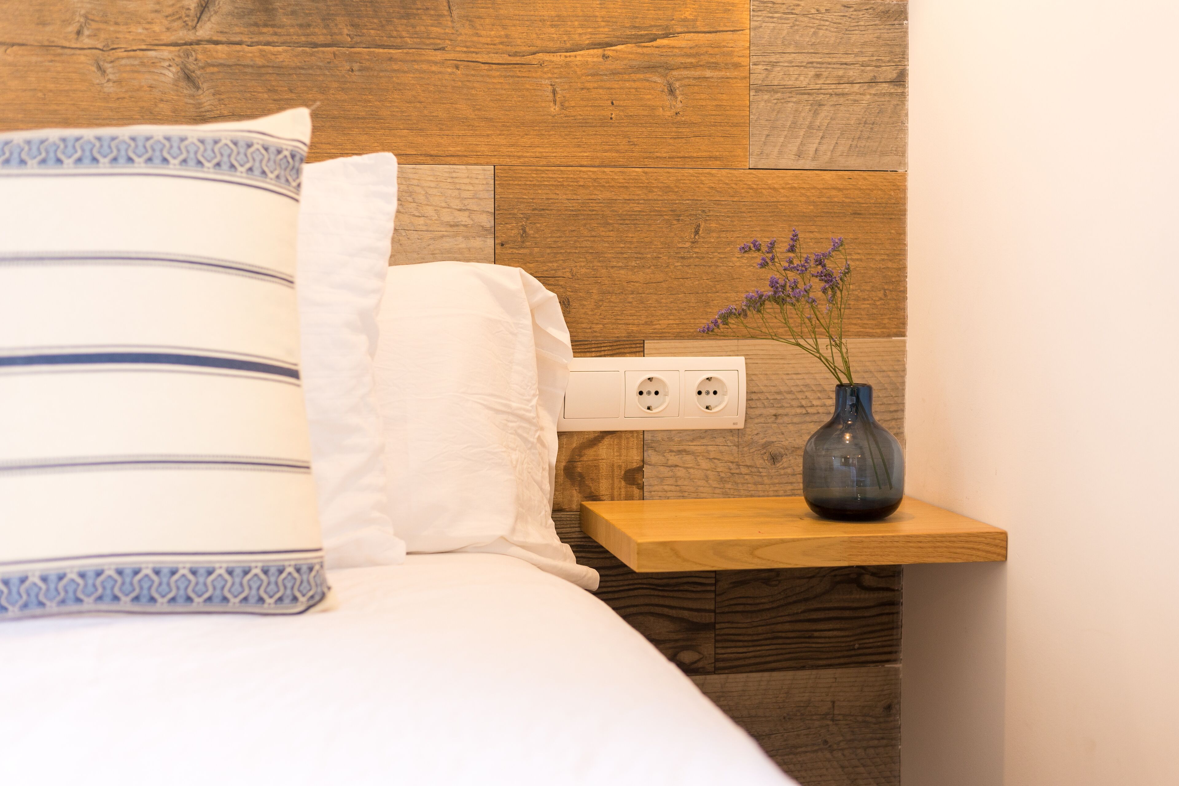 Una cama bien hecha con un cojín estampado, una mesita de noche con un jarrón de flores y un panel de pared de madera.
