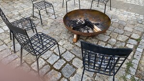 Un brasero rodeado por cuatro sillas de metal con asientos de madera, situado en un patio empedrado.
