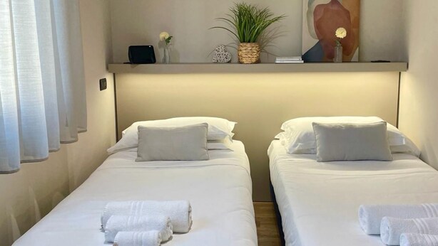 Une chambre d’hôtel avec deux lits simples, chacun muni d’un oreiller et d’une serviette roulée. Au-dessus des lits, on trouve une étagère ornée d’une plante, d’un objet décoratif et d’un tableau.