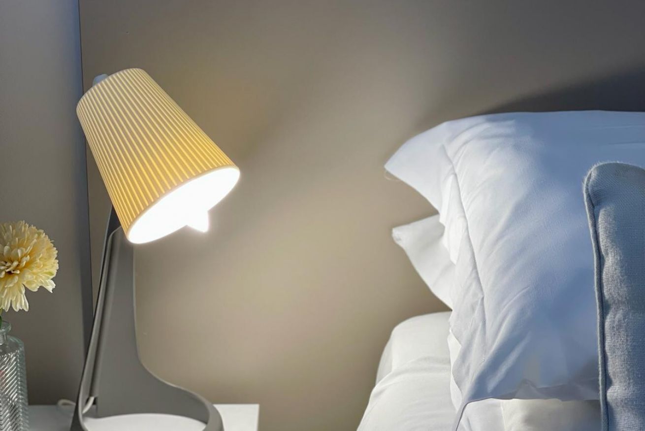 A bedside lamp with a ribbed lampshade on a nightstand next to a bed with white pillows and a vase with a yellow flower.