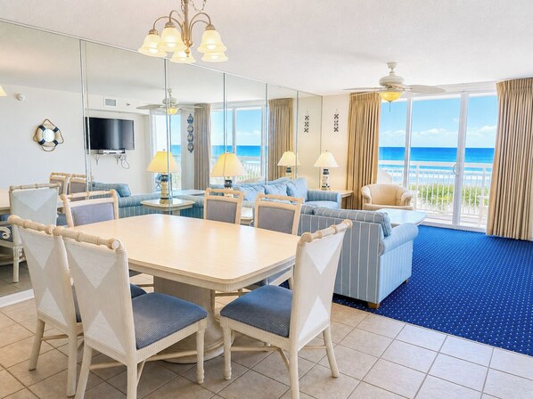A hotel room with a dining area, a sofa, a TV, and a balcony with a view of the ocean.