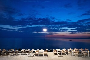 Eine Seeterrasse mit Tischen und Stühlen, eine Straßenlaterne und ein ruhiges Meer unter einem Zwielichthimmel.