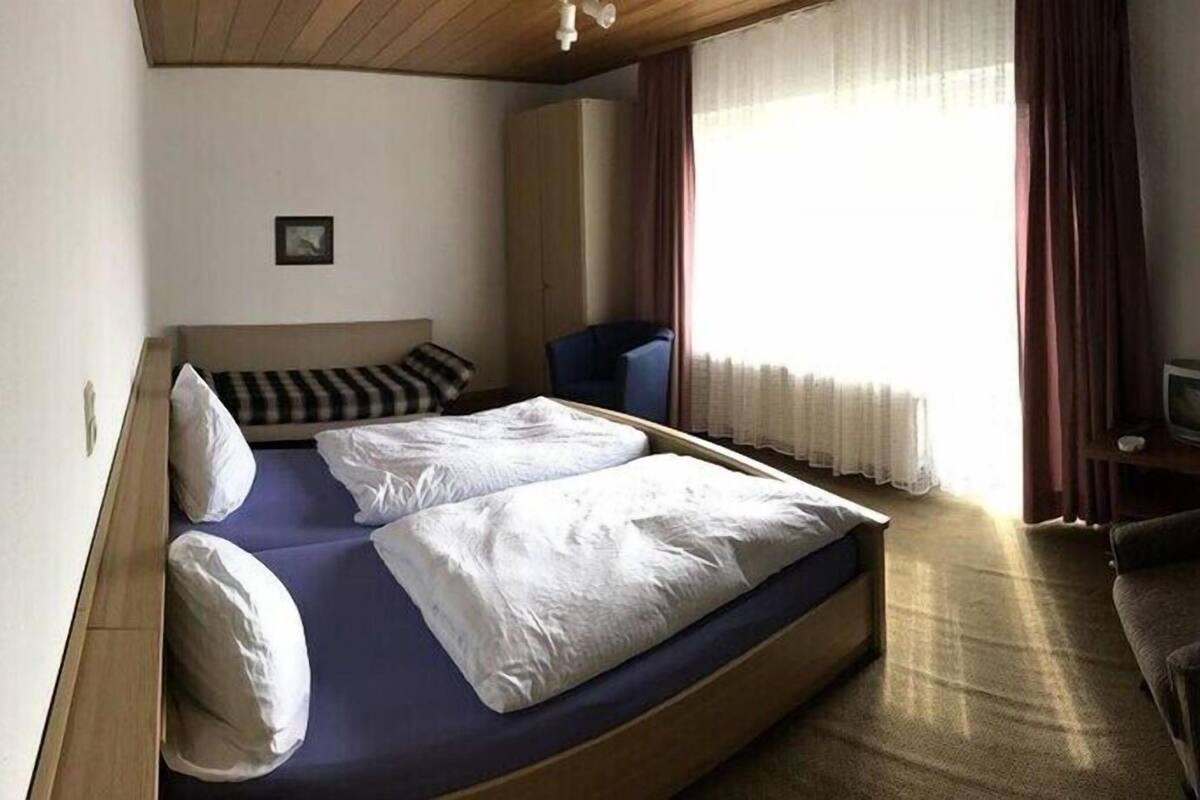 A small, compact hotel room with a single bed, a chair, a small table with a television, and a window with curtains.
