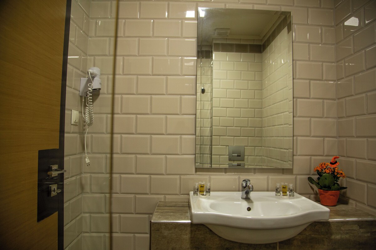 A bathroom with white tiled walls, a sink with a mirror above it, a hairdryer on the wall, and a potted plant with orange flowers.