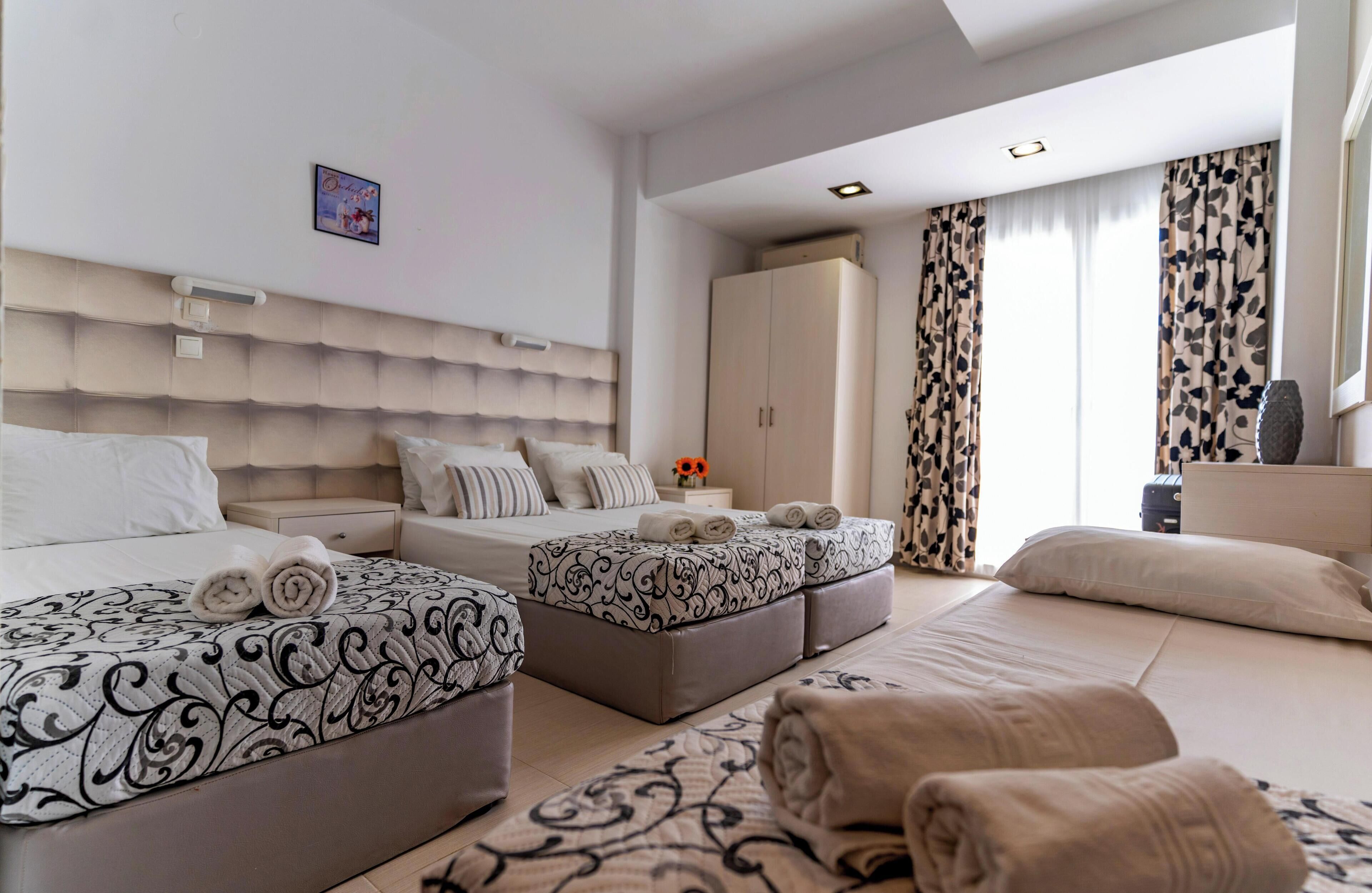 A hotel room with two beds, a wardrobe, a bedside table with a lamp, and a large window with curtains.