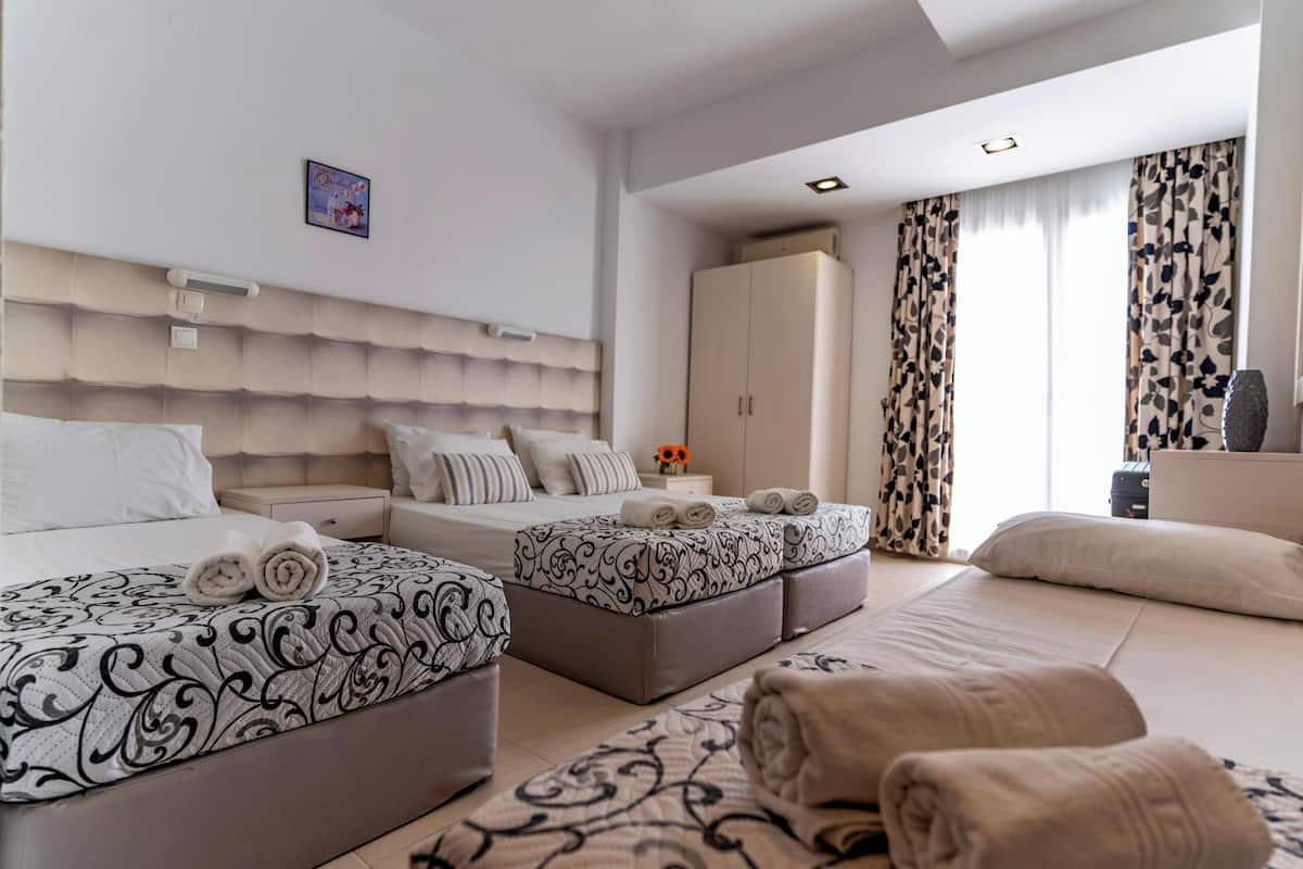 A hotel room with two beds, a wardrobe, a bedside table with a lamp, and a large window with curtains.