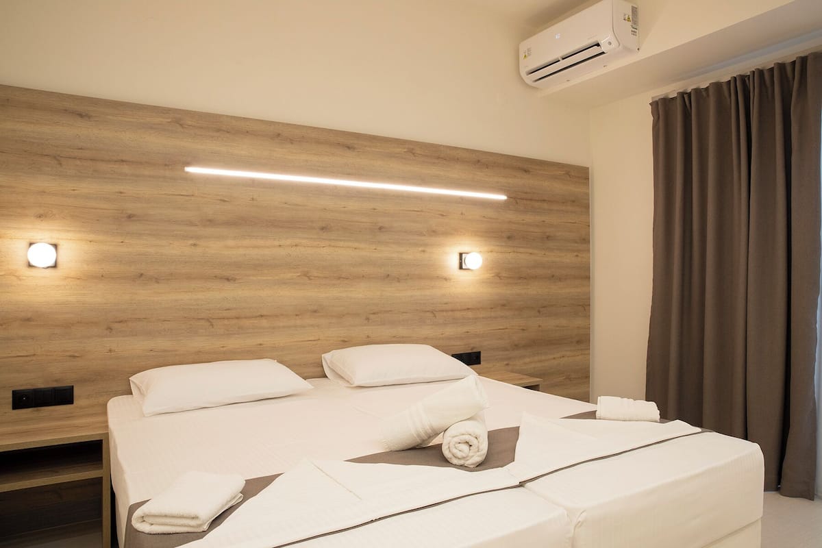 A hotel room with a wooden headboard, two bedside tables, a bed with white linens, a rolled towel, and an air conditioning unit.