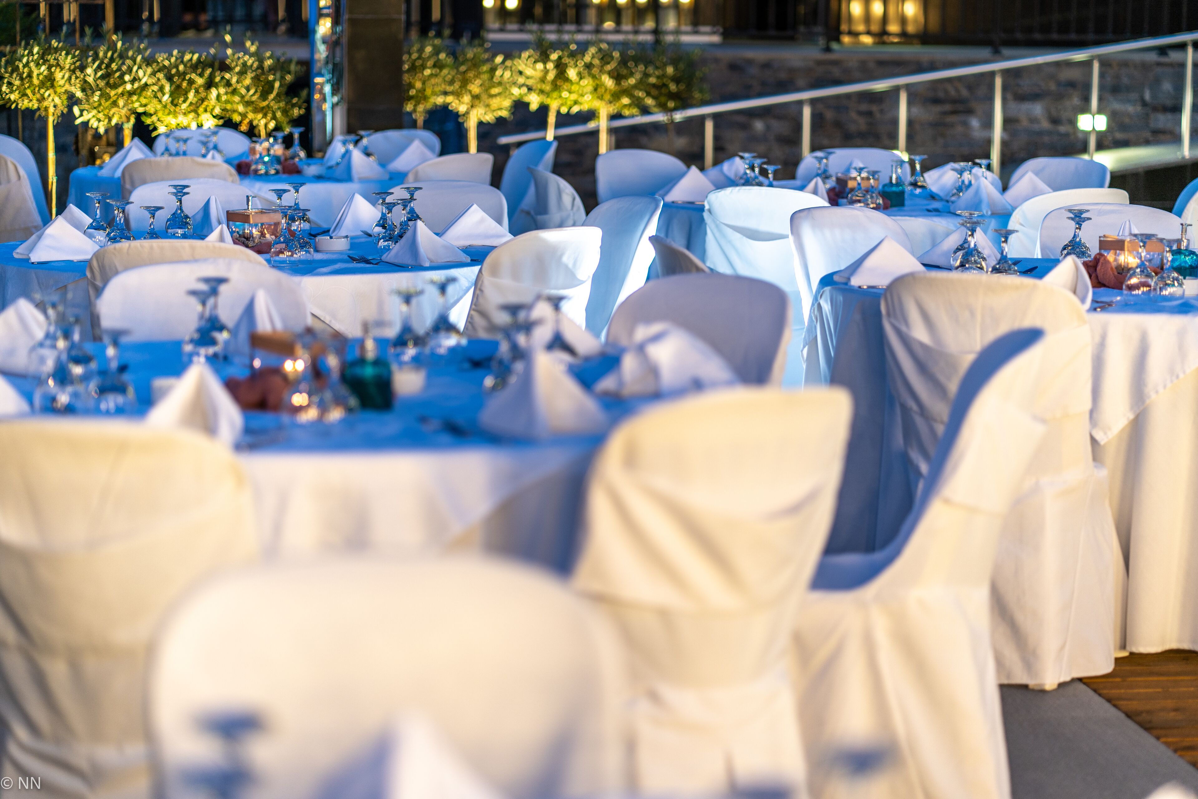 A formal outdoor dining area with tables set for a meal, surrounded by chairs with white and blue covers.