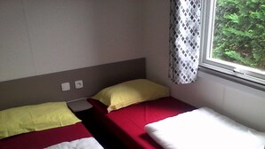 Una habitación pequeña con dos camas, cada una cubierta con una colcha roja y blanca. Se ve una ventana con cortina estampada, y las paredes son lisas.