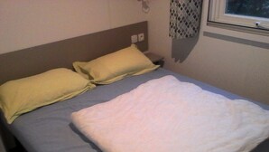 Una cama con una manta blanca y dos almohadas, una mesita de noche y una ventana con cortina.