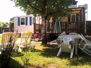 A backyard with a wooden deck, a white chair, a small table, and a tree. - Mobil Home Casita (Les Mathes)
