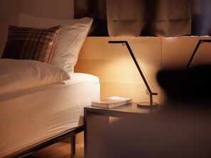 Una cama bien hecha con sábanas blancas y un cojín de cuadros, una mesita de noche con un libro y una lámpara, y una ventana con cortinas.