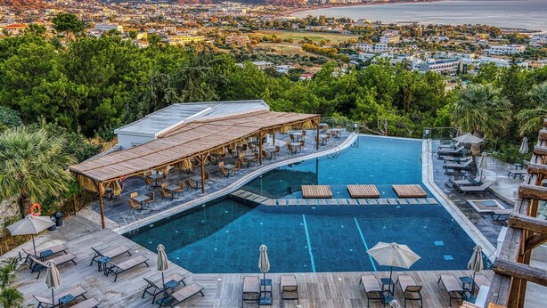 Piscina de resort con sillas de descanso y sombrillas, rodeada de vegetación exuberante y con vistas a una ciudad costera.