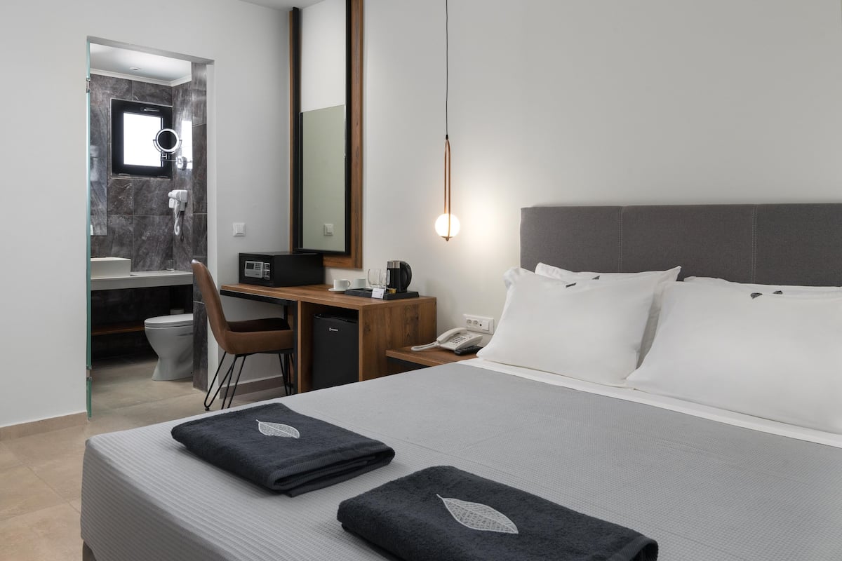 Une chambre d’hôtel avec un lit, un bureau, une chaise, un miroir, un téléphone et une salle de bain visible à travers une porte ouverte.