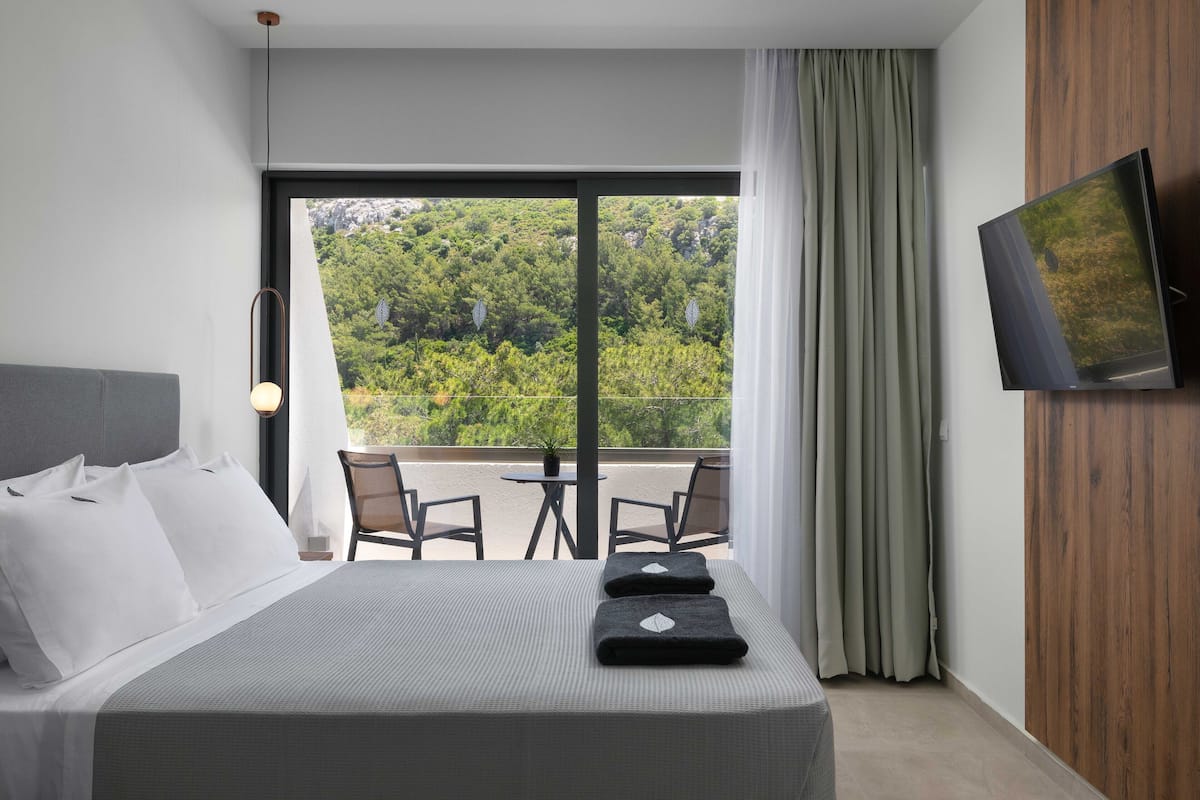 Une chambre d’hôtel avec un grand lit, un téléviseur fixé au mur, un balcon donnant sur un espace verdoyant et une table avec des chaises.