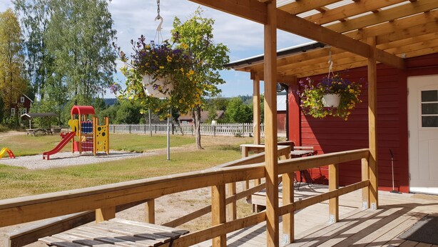 A wooden deck with a red building in the background, a playground with slides, and hanging flower baskets.