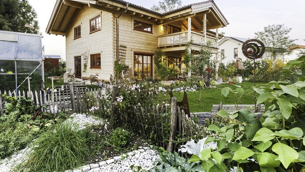 A two-story house with a balcony, surrounded by a garden with various plants and a decorative metal sculpture. - Fantastic apartment in a log cabin in front of the mountains (Wielenbach)