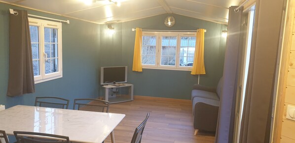 A room with a dining table, chairs, a television on a stand, and a window with curtains. - Camping de La Pointe (Bourg-et-Comin)