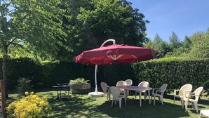 A patio with a table and chairs under a red umbrella, surrounded by greenery and a hedge.