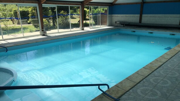 An indoor swimming pool with clear blue water, a tiled floor, and a covered area with large windows.