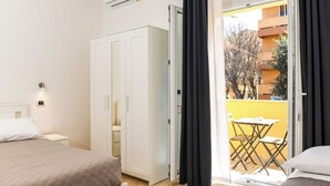 Een hotelkamer met een bed, een kledingkast, een klein tafeltje met stoelen en een balkon met een gele muur en bomen.