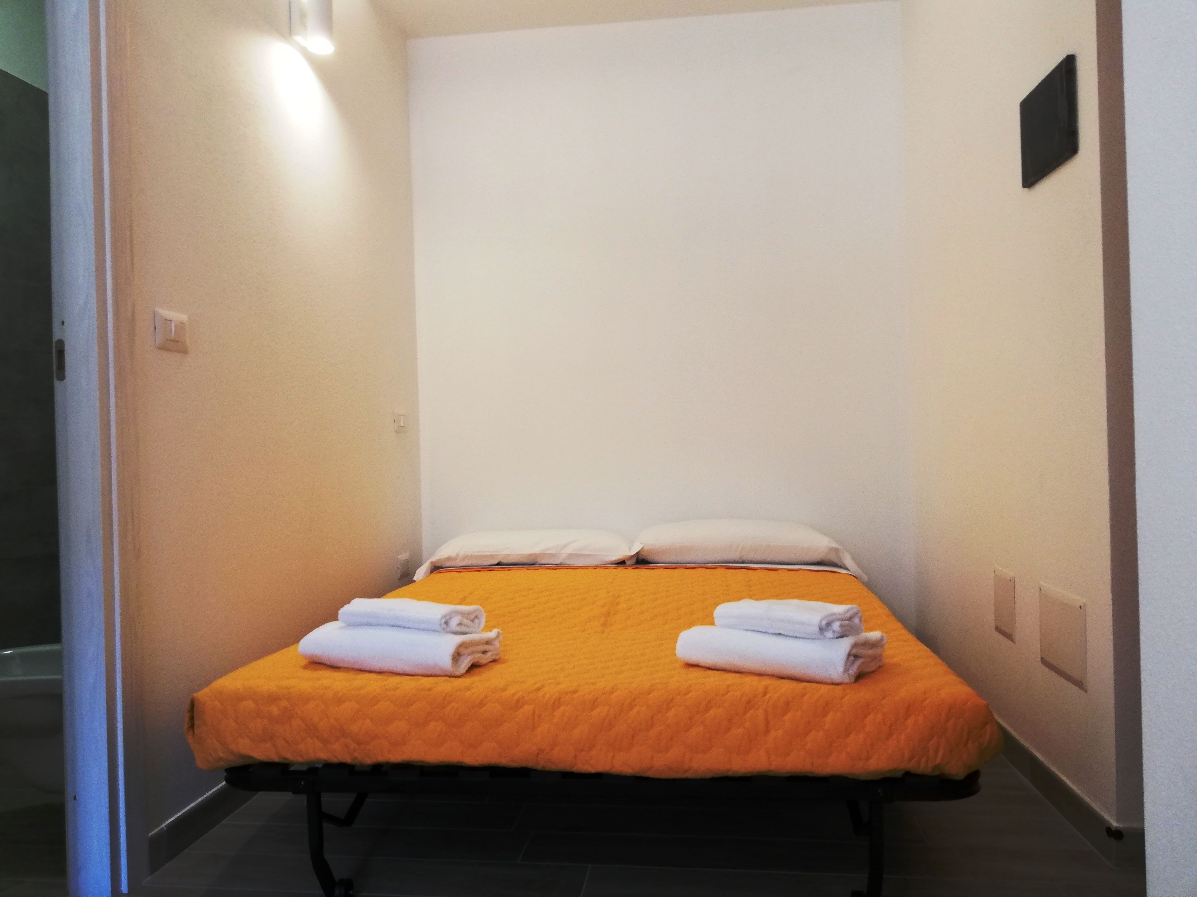 Ein kleines Zimmer mit einem Einzelbett, einer orangefarbenen Bettdecke, zwei weißen Kissen und zwei ordentlich gefalteten Handtüchern.