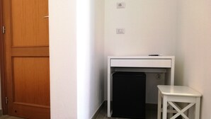 Een kleine kamer met een houten deur, een wit bureau met een zwarte computer en een kleine witte kruk.