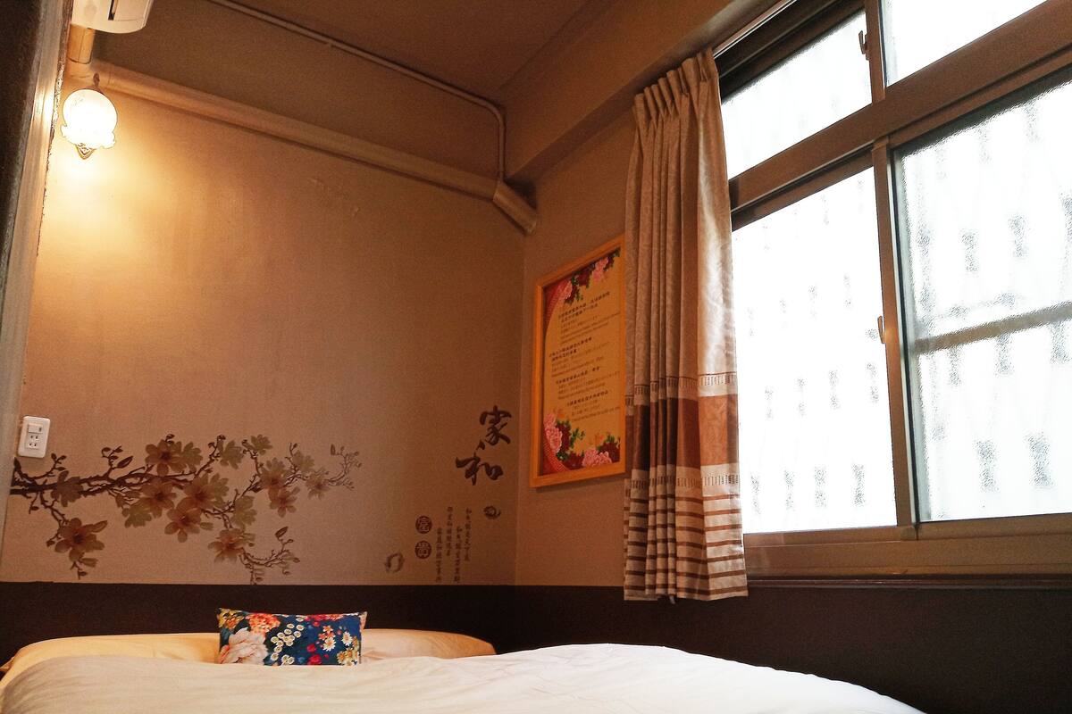 A hotel room with a bed, a wall-mounted light, a window with curtains, and a decorative wall piece.