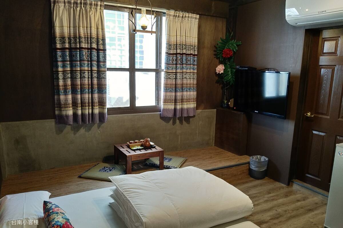 A room with a bed, a small table with a teapot, a TV, and a window with curtains.