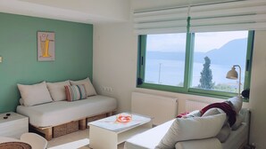 A cozy room with a sofa, a small table, a window with a view of the sea, and a bed in the background.