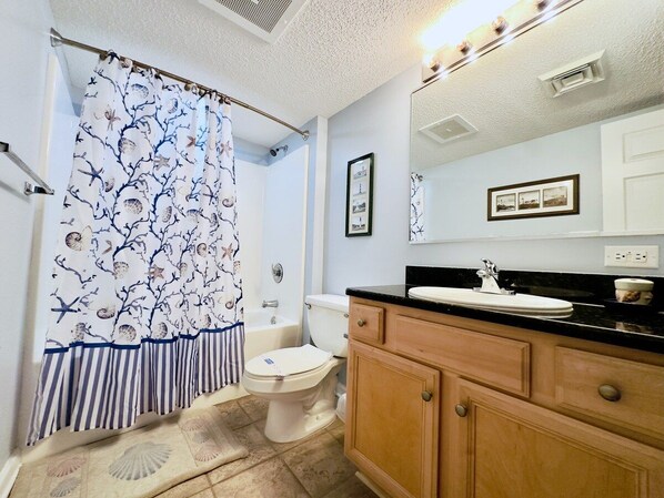 A bathroom with a white toilet, a wooden vanity with a black countertop, a shower curtain with a nature pattern, and a mirror above the vanity.