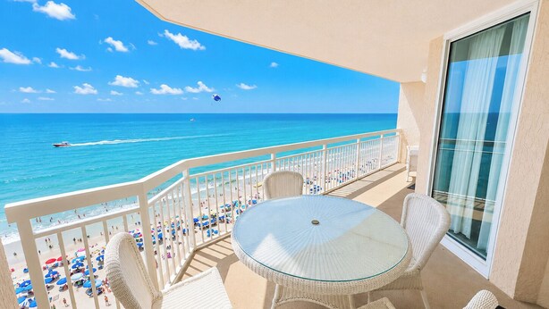 A balcony with a glass table and wicker chairs, overlooking a beach with umbrellas and people. - Crescent Shores S 1309 - Oceanfront Luxury & Unmatched Views! (North Myrtle Beach)