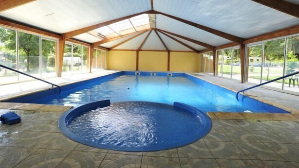 Una piscina interior con suelo de baldosas, vigas de madera y grandes ventanales.