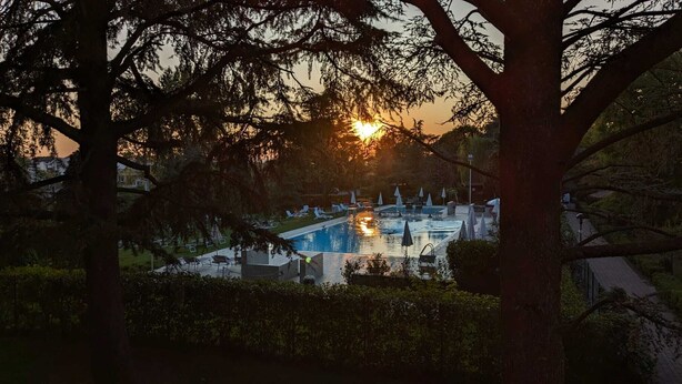 A swimming pool area with lounge chairs and umbrellas, surrounded by trees and a sunset in the background. -  TWO-ROOM APARTMENT SURROUNDED BY GREEN IN A QUIET RESIDENCE WITH POOL (Sirmione)