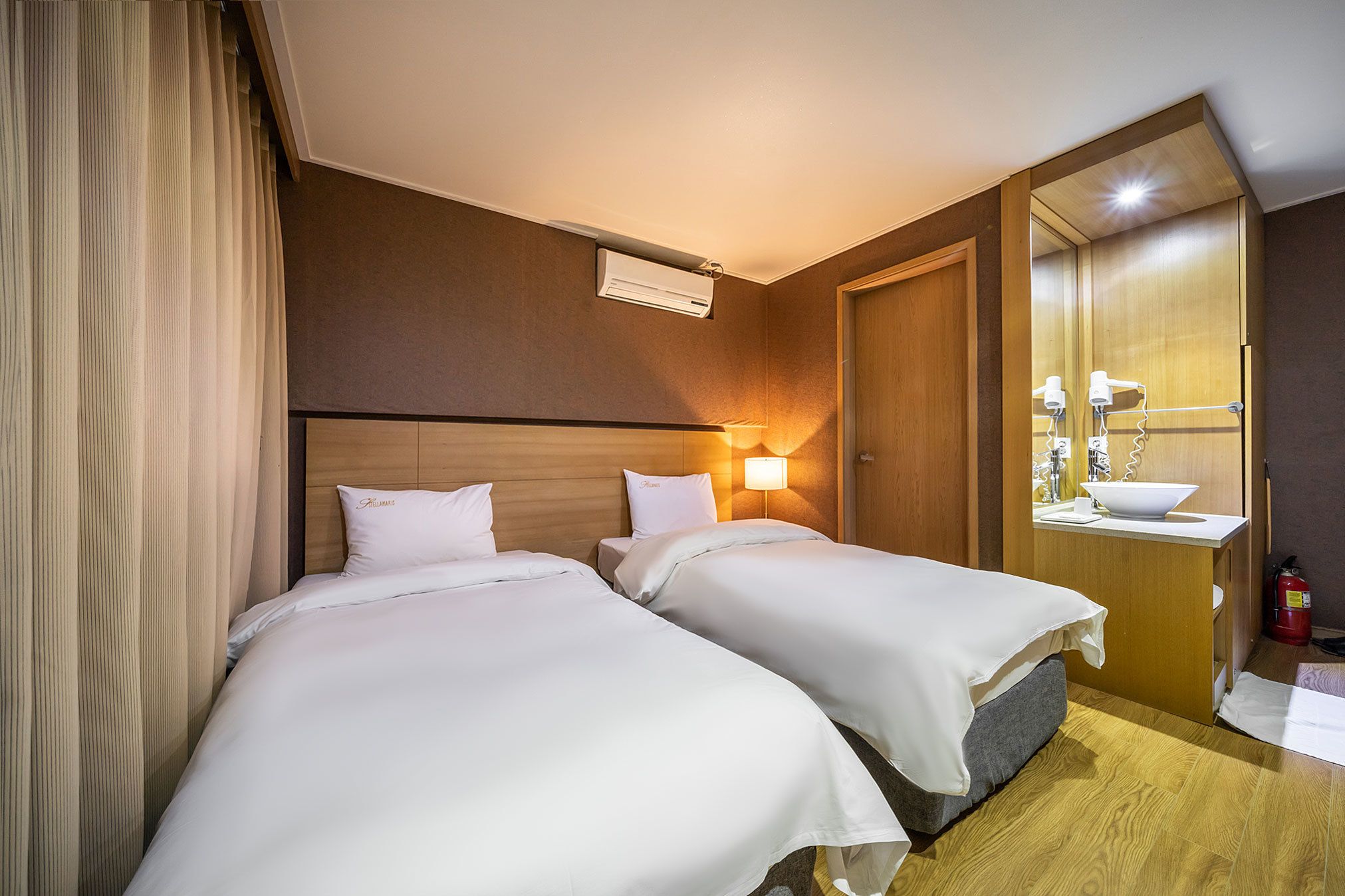 A hotel room with two beds, a wooden headboard, a bedside lamp, a mirror, and a small bathroom with a sink and towel rack.