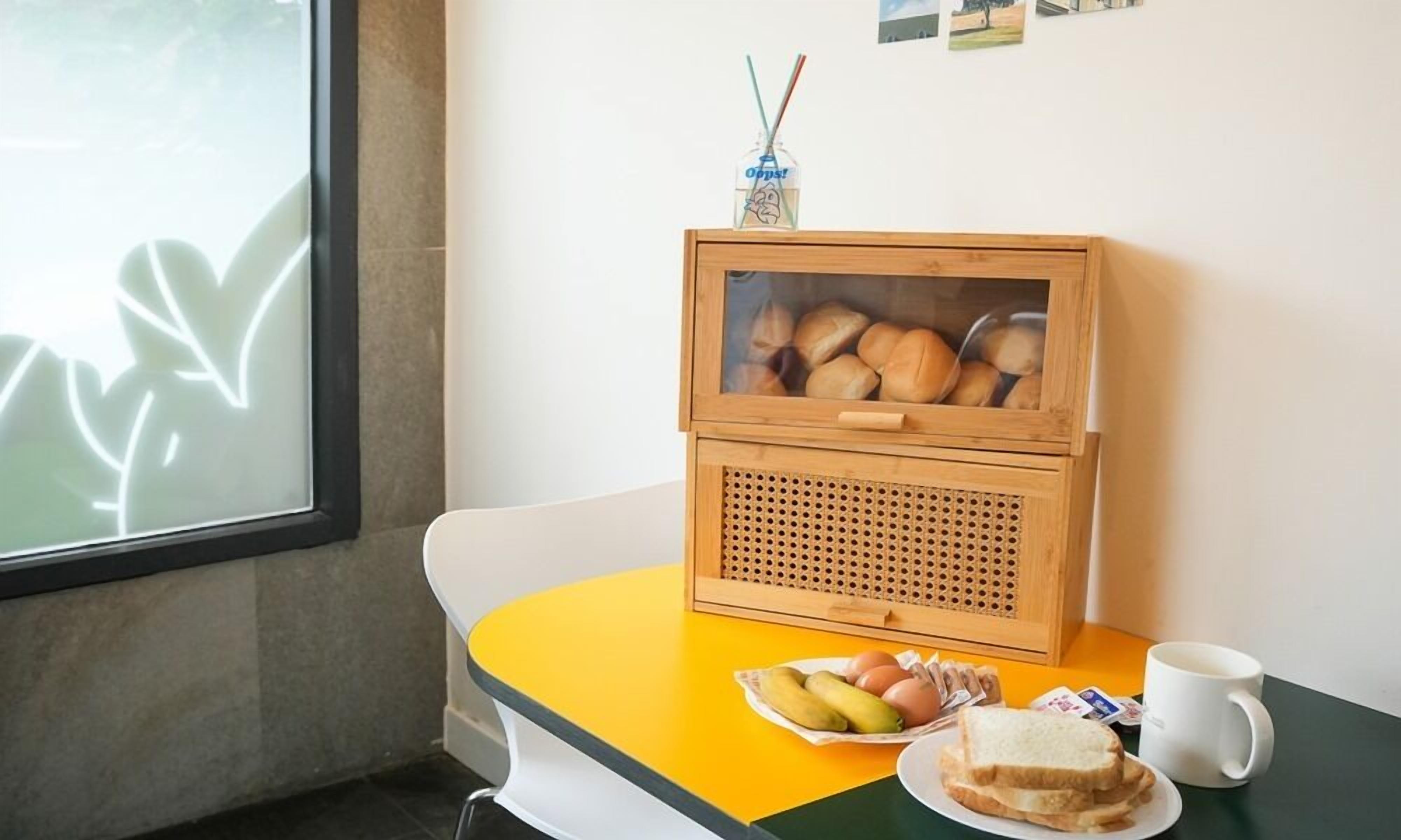 A small wooden cabinet with a glass front containing bread, a tray with eggs, bananas, and a plate with sandwiches, and a white mug on a yellow table.