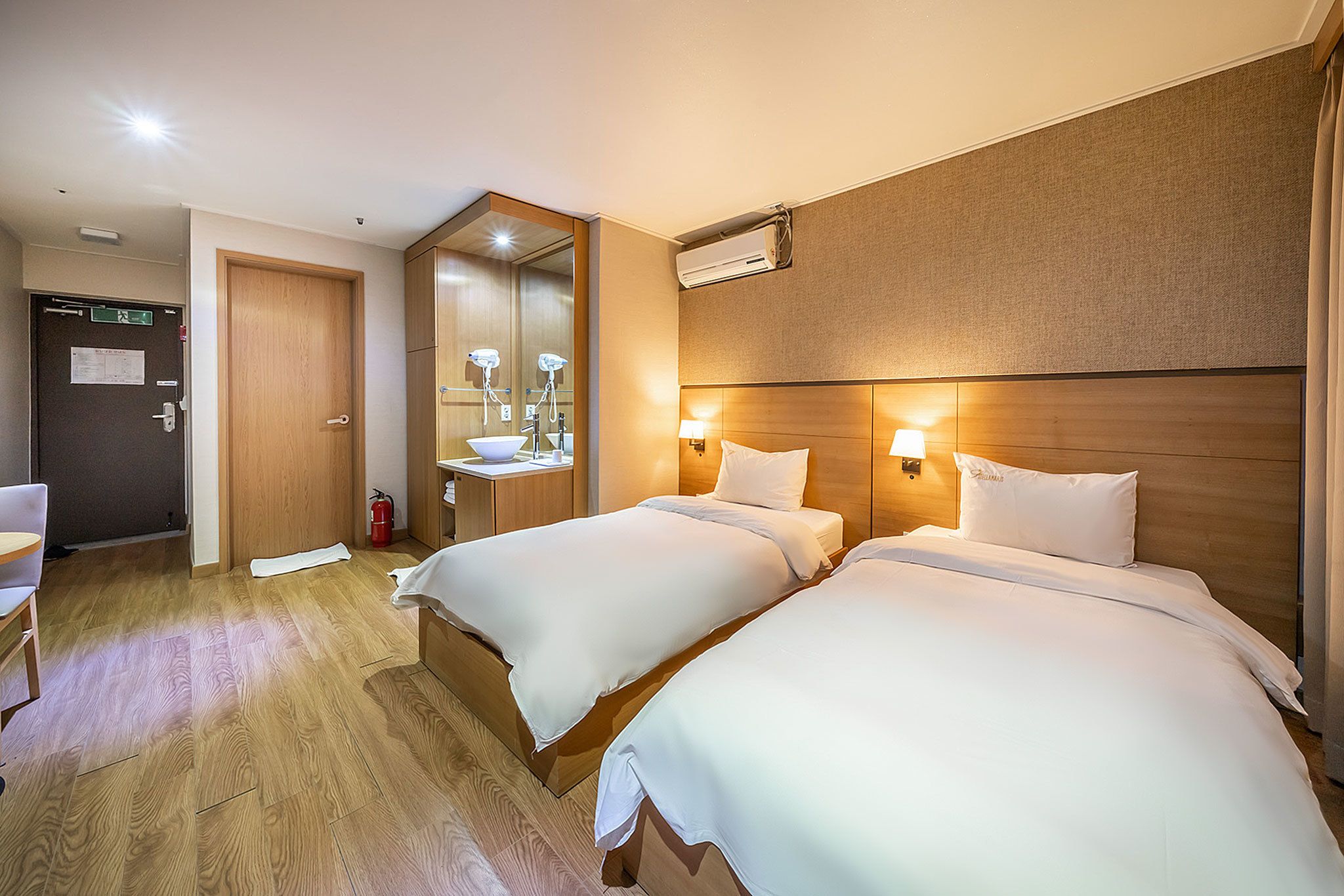 A hotel room with two beds, a wooden headboard, a mirror, a sink, and a chair.