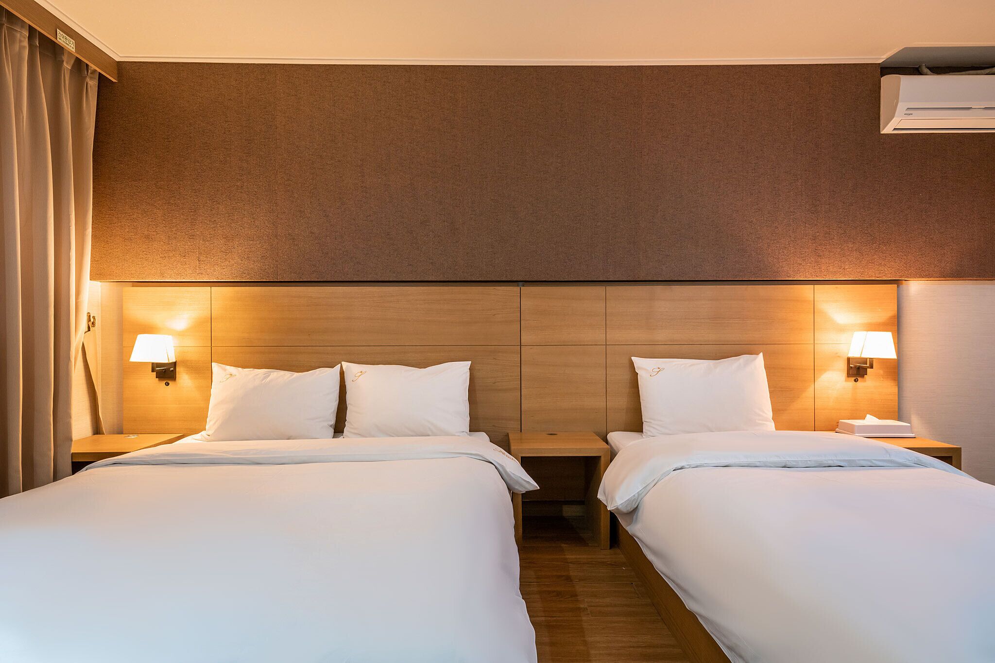 Ein Hotelzimmer mit zwei Betten, Holzkopfteilen, Wandleuchten und einer Klimaanlage.