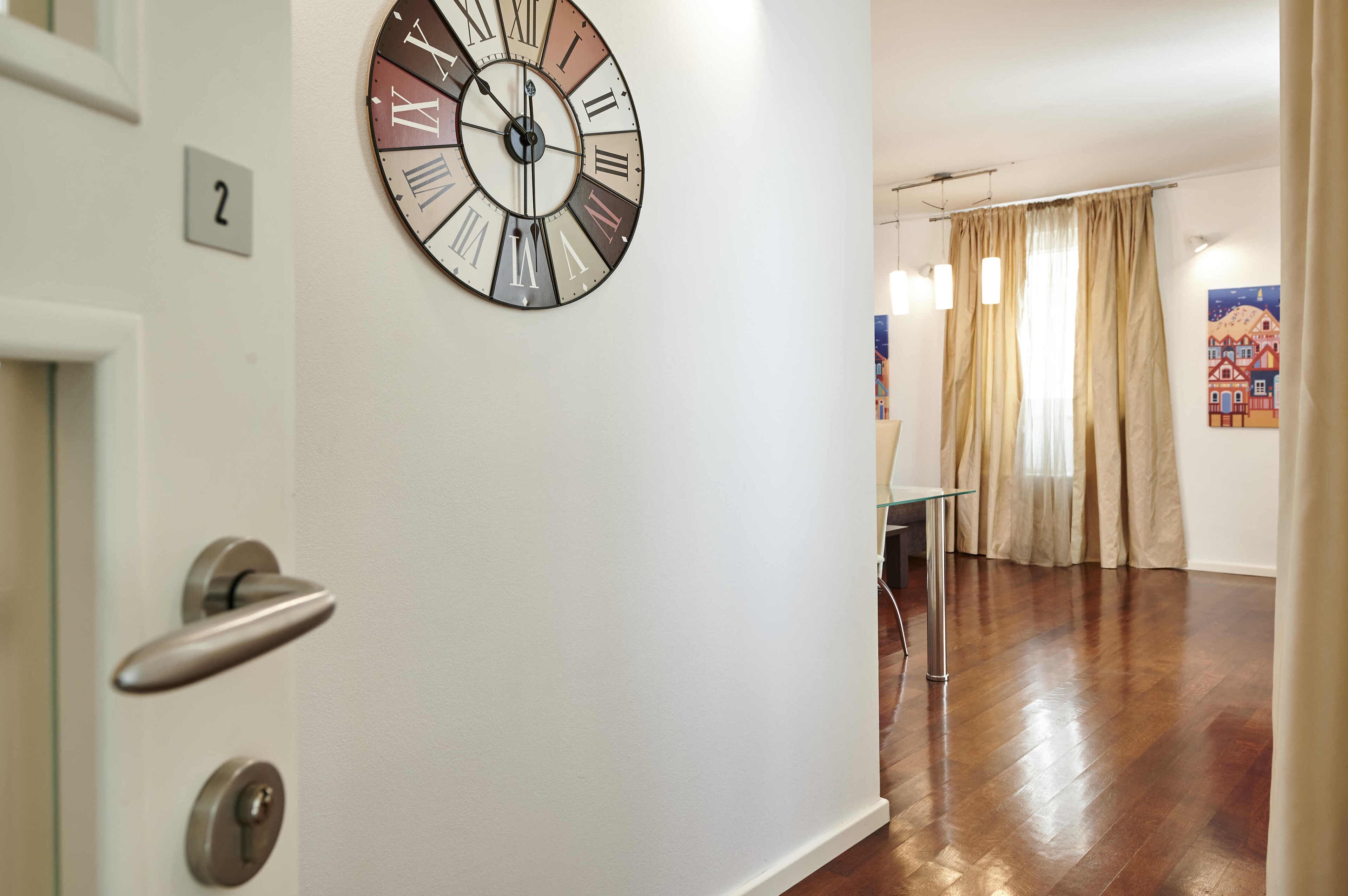 A room with a large wall clock, a door with a number plate, and a window with curtains.