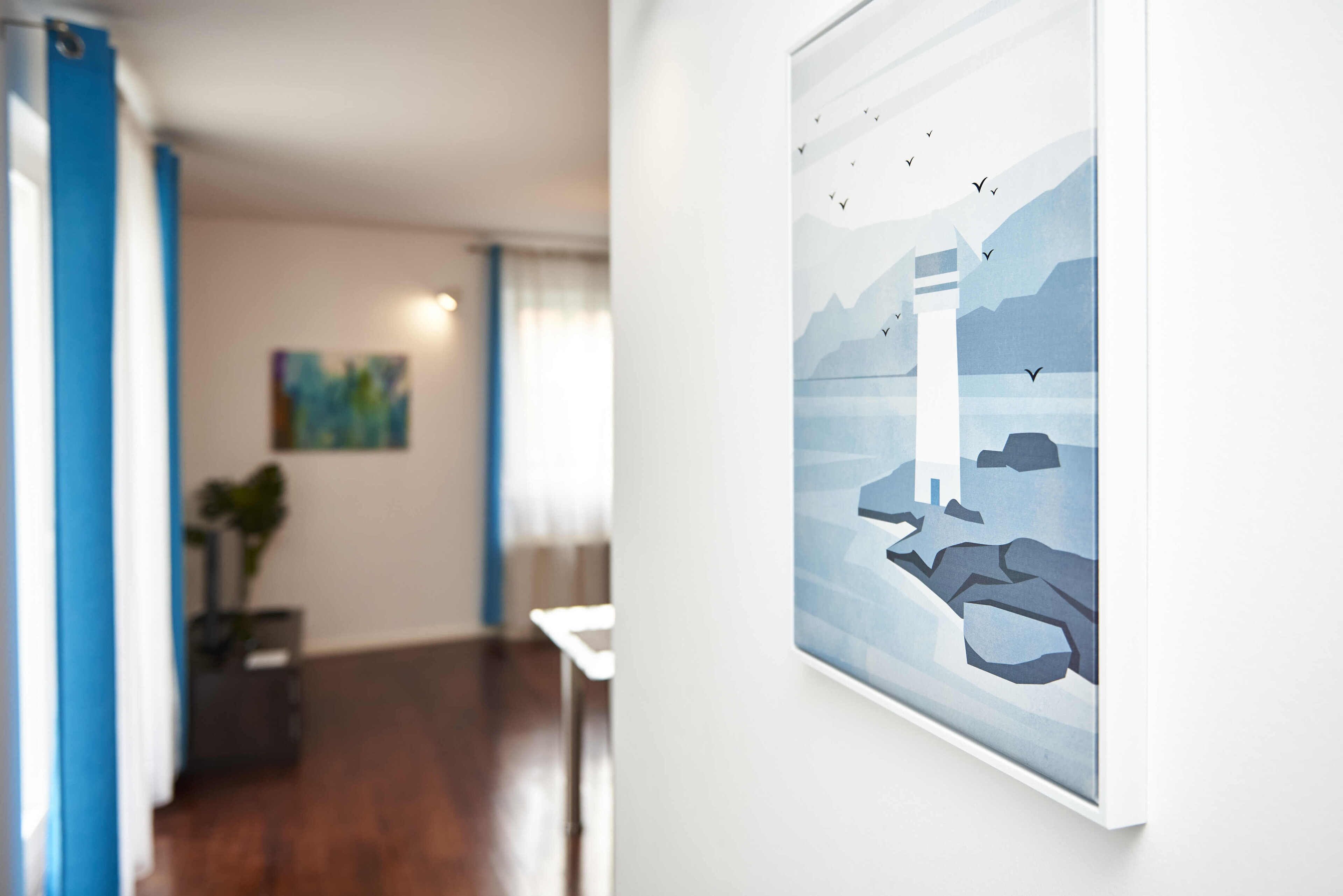 A hallway with a framed artwork depicting a lighthouse and birds, a potted plant, and a painting on the wall.