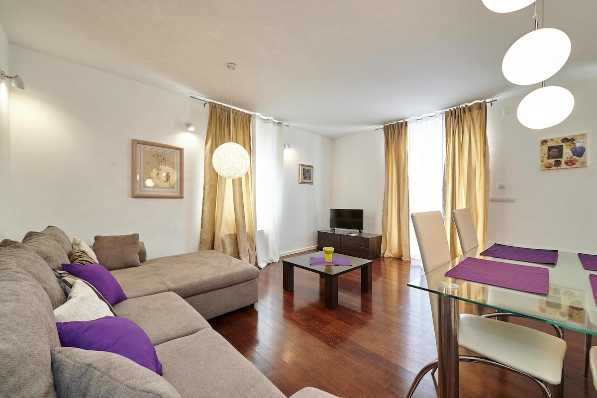 Uma sala de estar com sofá, mesa de centro de vidro, televisão e uma área de jantar com cadeiras e mesa.