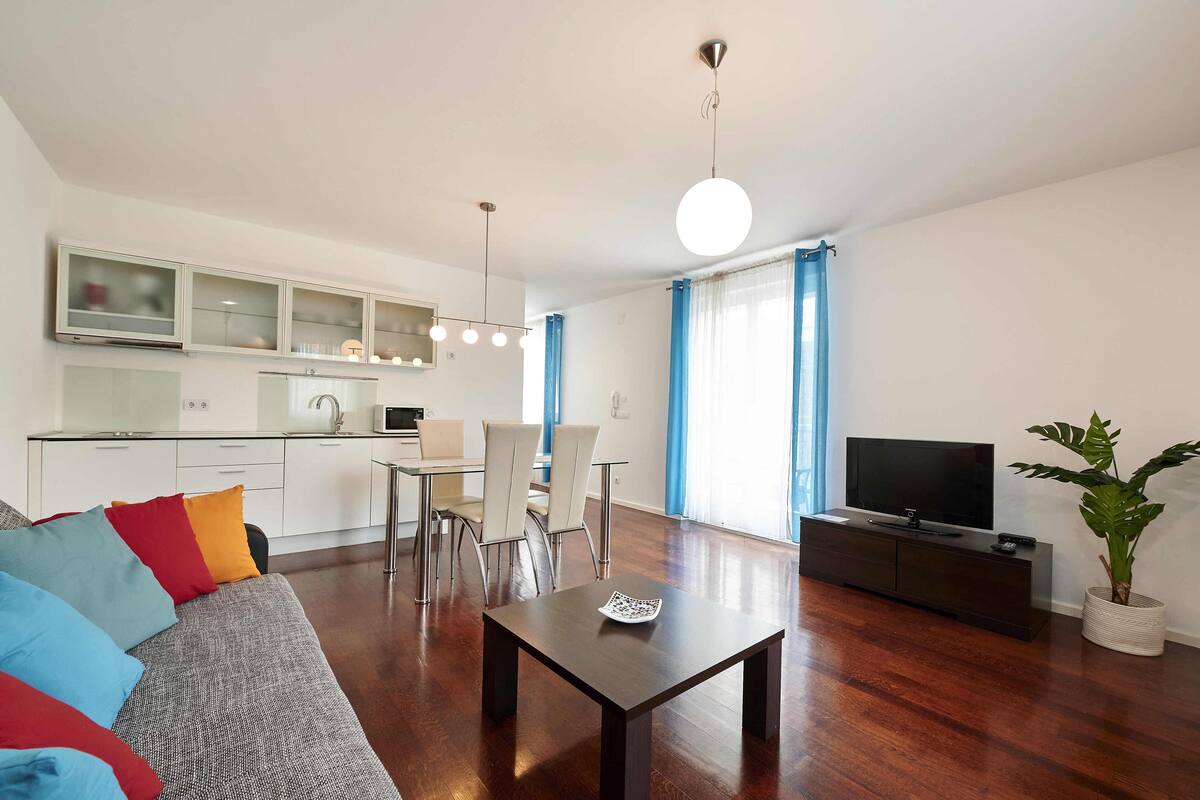 Sala de estar moderna com sofá, TV, mesa de jantar e área de cozinha ao fundo.