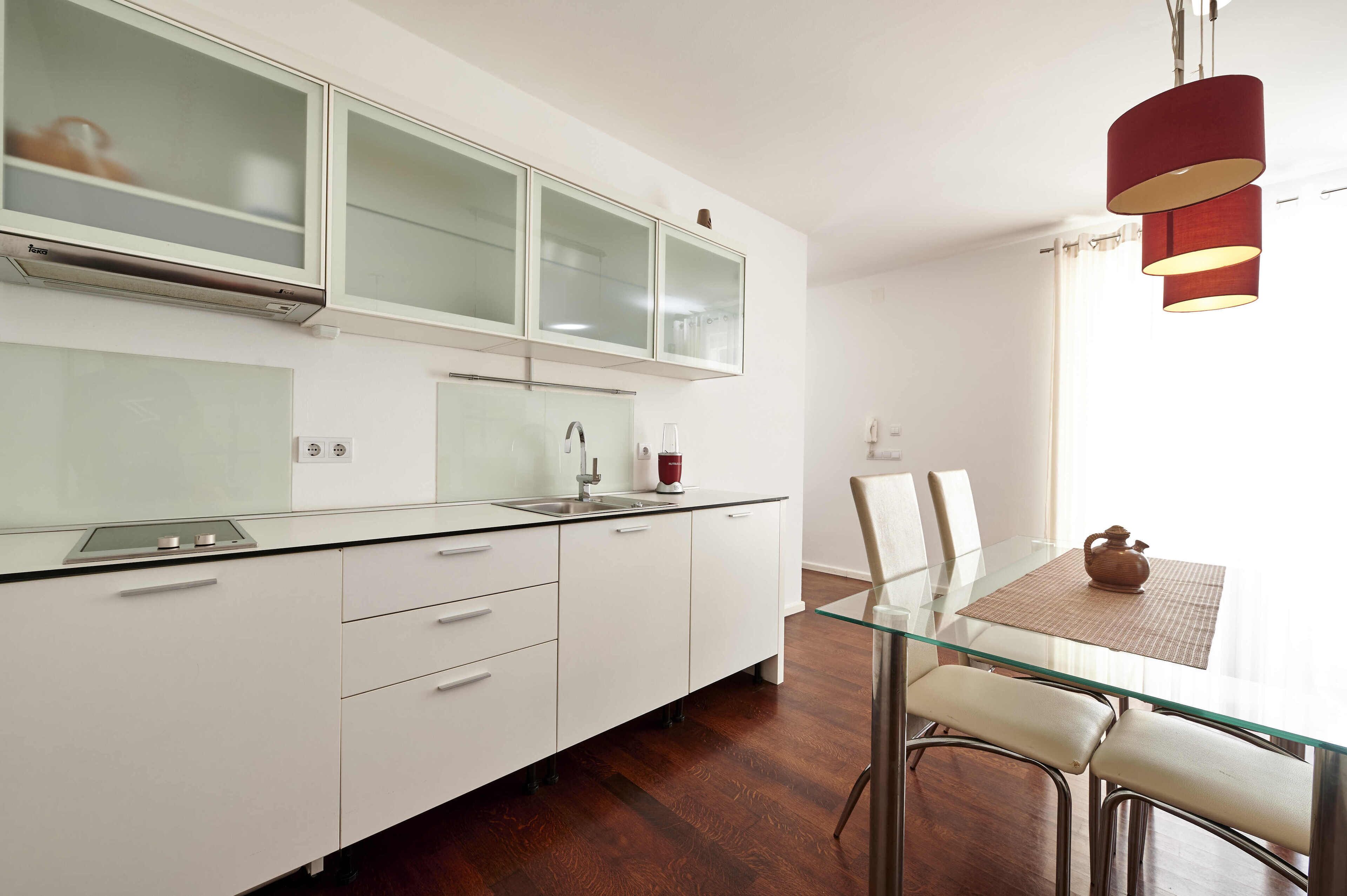 Una cocina moderna con armarios blancos, una mesa de comedor con cubierta de vidrio y una lámpara colgante roja.