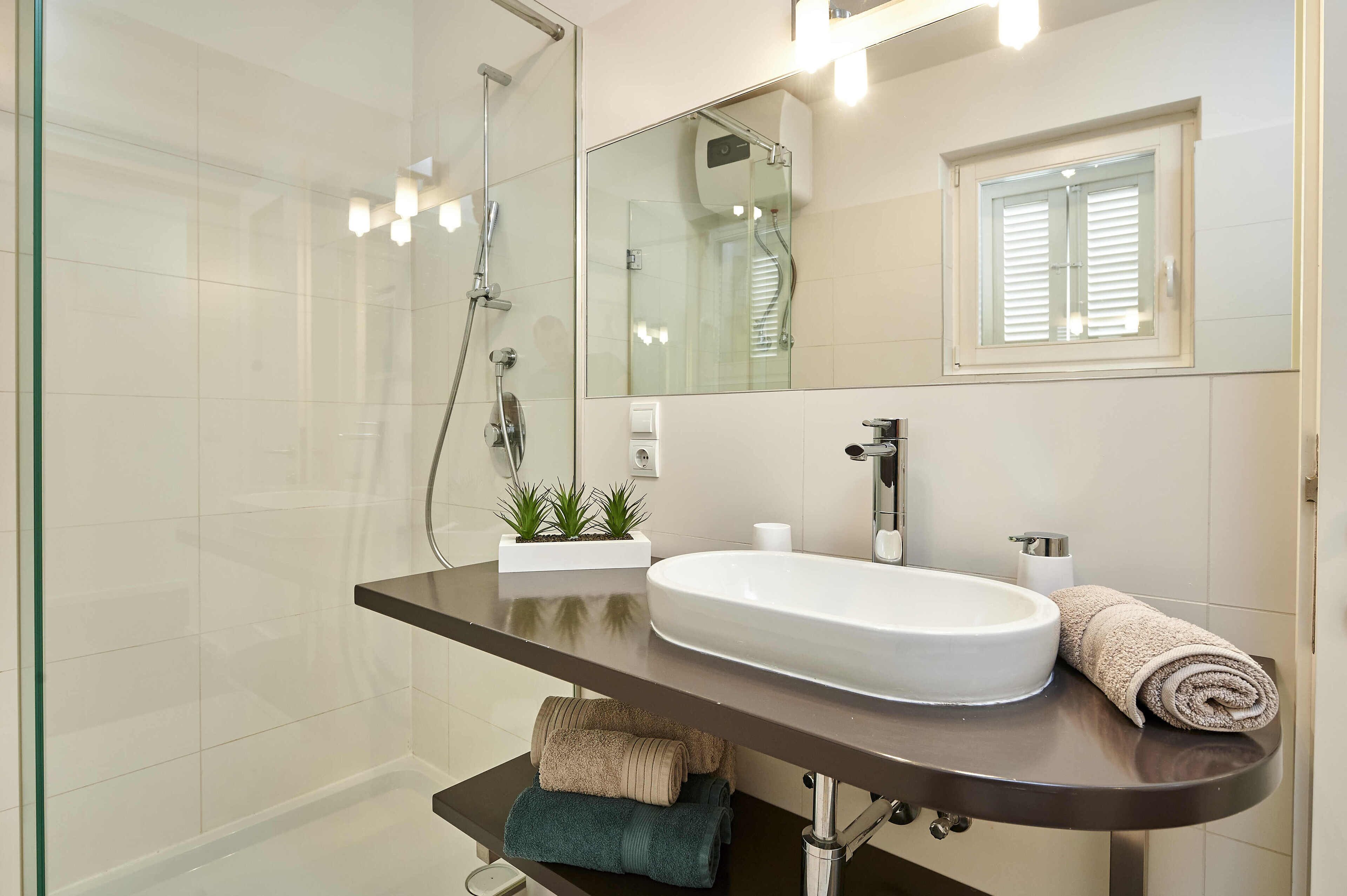 A modern bathroom with a glass shower enclosure, a large mirror, a white sink, and a window with blinds.