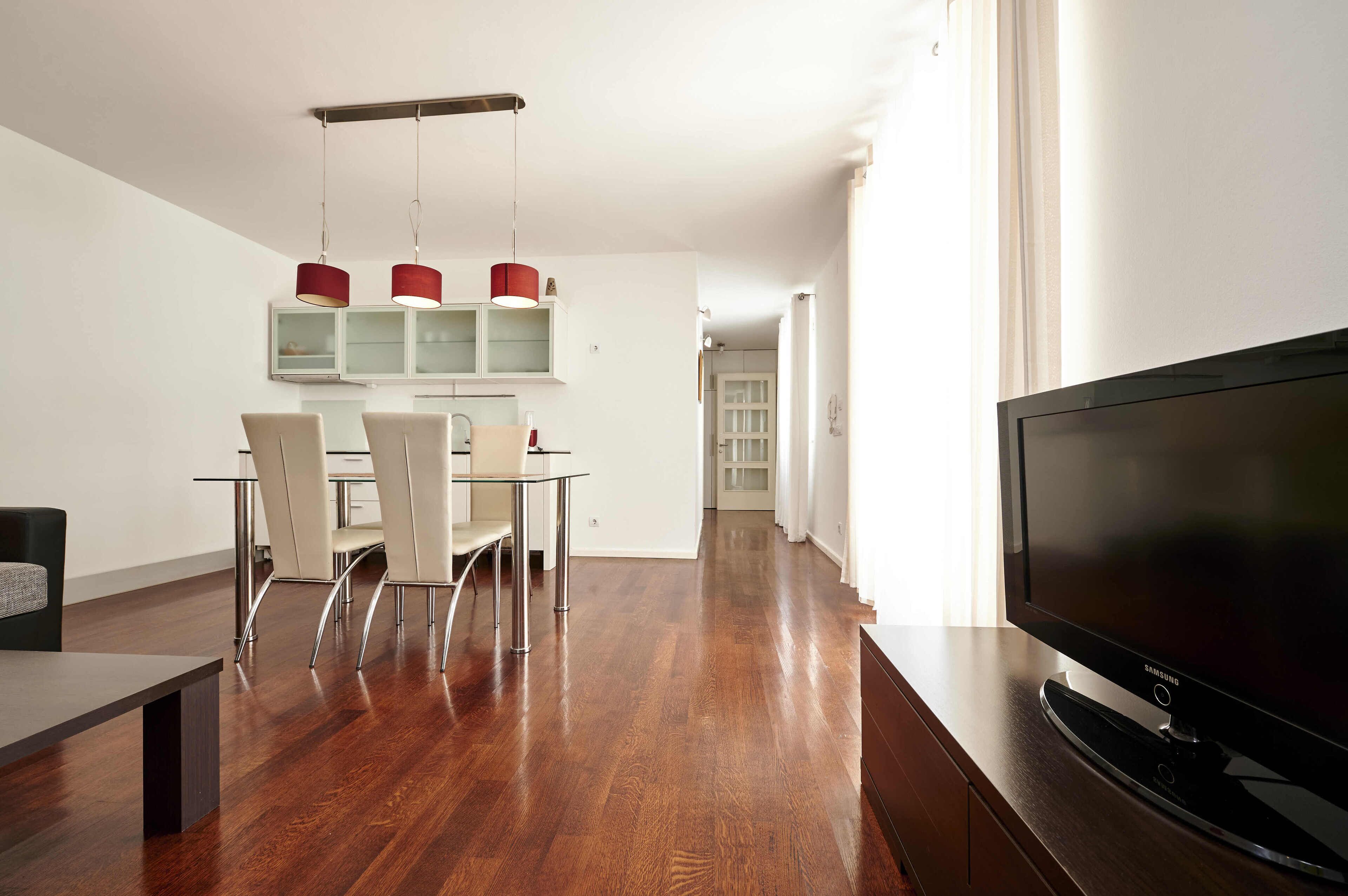 A modern living room with a flat-screen TV, a dining area with a glass table and chairs, and a kitchen with red pendant lights.